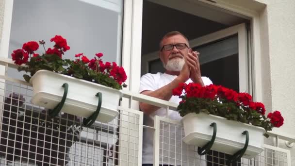 Graubärtiger alter Mann, der auf seinem Balkon applaudiert, um medizinisches Personal während der globalen Pandemie zu unterstützen. Soziale Distanz und Selbstisolierung — Stockvideo