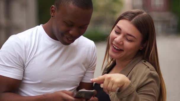 Міжрасова пара розважається зі смартфоном на міській вулиці. Концепція молодого способу життя. Чорний чоловік і кавказька жінка сміються і дивляться фотографії в смартфоні. Пара перегонів. — стокове відео