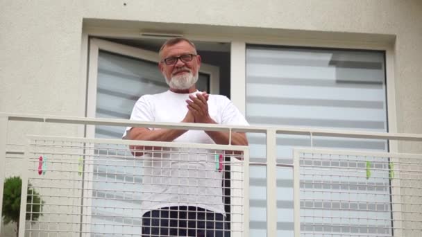 Pensionné avec des lunettes se tient sur le balcon et applaudit ses mains en remerciement aux médecins pour leur travail. Les médecins soutiennent l'action pendant la quarantaine du coronavirus covid-19 — Video