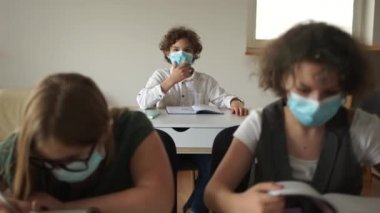 Kıvırcık maskeli okul çocuğu cevap vermek için elini kaldırdı. Coronavirus covid-19 karantinasından sonra sınıftaki çocuklar karantina sonrası okula dönüyorlar.
