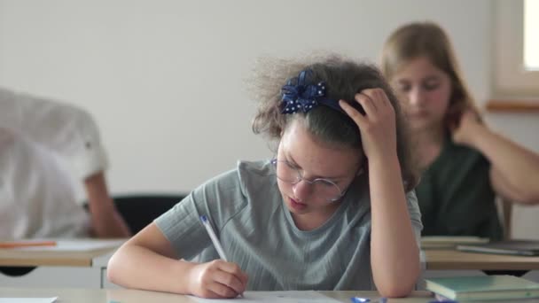 Nahaufnahme eines süßen Schulmädchens mit lockigem Haar und Brille. Mädchen schreibt im Unterricht in Notizbuch — Stockvideo