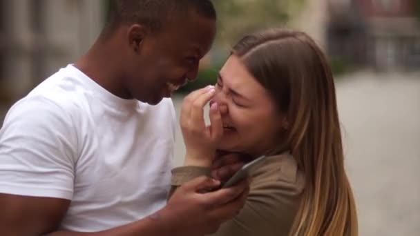 Gemengde race koppel date. Interraciaal stel heeft plezier met smartphone in de stad straat. Jonge levensstijl concept. Een zwarte man en een blanke vrouw lachen en kijken foto 's in een smartphone — Stockvideo