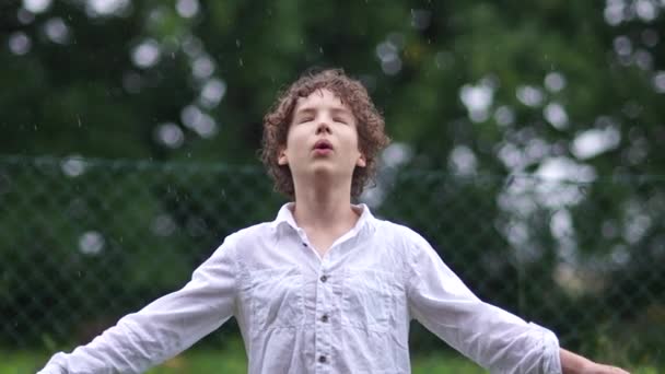 Retrato de niño lindo que se divierte atrapando gotas de lluvia. Un colegial de pelo rizado con una camisa blanca extendió los brazos para encontrarse con la lluvia. Lluvia cálida de verano, estación húmeda, tiempo lluvioso — Vídeos de Stock