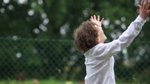 Kinky écolier se réjouit de la pluie d'été. Un garçon en chemise blanche encercle ses bras tendus sous des gouttes de pluie. Prévisions météorologiques, enfance heureuse — Video