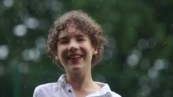 Крупним планом портрет симпатичного молодого хлопчика, який весело ловить краплі дощу. Щасливий підліток дитинства у виливний дощ, вологий сезон — стокове відео