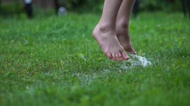 裸足のティーンエイジャーのクローズアップスローモーションショットは、水がスプリンクルしながら草の中にジャンプします。泥だらけの水たまりの人々。濡れた緑の草の上を歩く男性の足。幸せな子供時代のコンセプト. — ストック動画