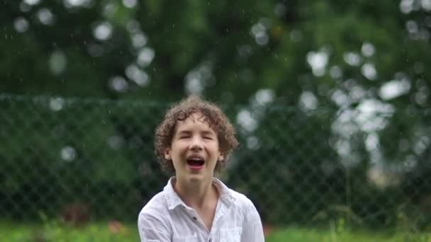 Glückliche verrückte lockige Teenager springen und Spaß im Park im Regen haben. Ein Schuljunge im weißen Hemd jubelt im warmen Regen. Glückliche Kindheit — Stockvideo