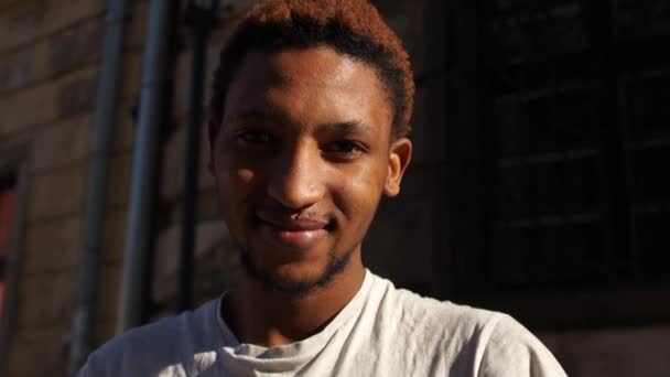 Czarny uśmiecha się patrząc w kamerę. Portret młodego Afroamerykanina w białej koszulce na ulicy. Pomysł szczęśliwych ludzi, stop rasicm — Wideo stockowe