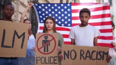 Farklı ten renginde genç öğrenciler, Amerikan bayrağının arka planına karşı ırkçılığı ve şiddeti protesto ediyorlar. Genç oğlanlar ve kızlar pankart tutuyorlar. Irkçılık yok, BLM, polis yasak bölge.