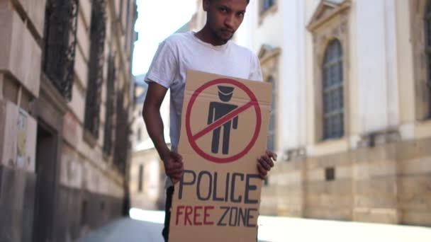 Un estudiante negro con una camiseta blanca está en medio de una calle de la ciudad. Hombre afroamericano sosteniendo un cartel con la inscripción POLISE FREE ZONE. Protestas contra la violencia policial — Vídeo de stock