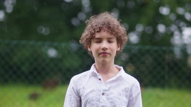 Schulkind hat Spaß im warmen Sommerregen. Nahporträt lockiger Junge im weißen Hemd, glückliche Kindheit — Stockvideo