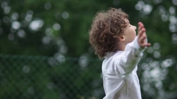 Ein fröhlicher Schuljunge im weißen Hemd amüsiert sich im strömenden Regen. Warmer Sommerregen, glückliche Kindheit — Stockvideo