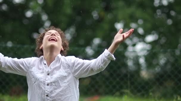 Rapaz está à chuva e ri com os braços afastados. Adolescente em uma camisa branca durante uma tempestade. Fechar retrato de um estudante de férias — Vídeo de Stock