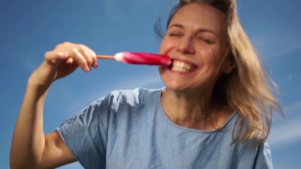 Gelukkige vrouw met een witte glimlach eet rood ijs op een stok op een hemelse achtergrond. Reisconcept — Stockvideo