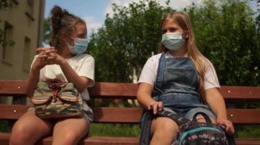 Maskeli iki güzel kız sırt çantalı bir bankta oturuyorlar. Elveda ulaşılamaz, sosyal uzaklık, okula dönüş, karantina hayatı