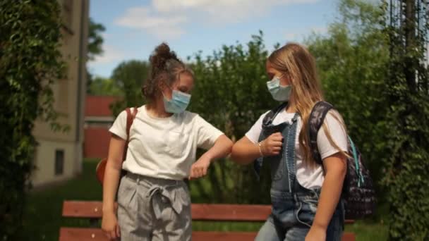 Két gyönyörű lány, maszkos tinédzserek köszöntik a kapcsolat hiányát, a társadalmi távolságot, vissza az iskolába, fazekas-karantén életet. — Stock videók