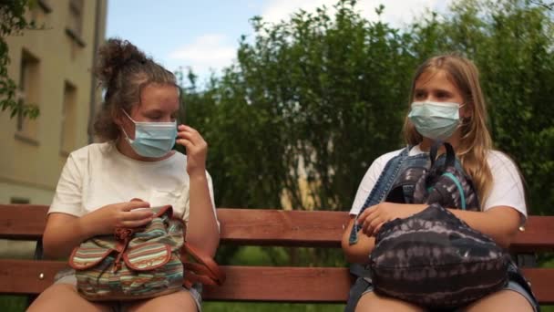 Schoolgaande kinderen twee meisjes met medische maskers zitten op een bankje. Het meisje deed haar masker af en ademt zwaar. Zuurstofverhongering. Pandemische coronovirus covid-19. Het leven na de quarantaine — Stockvideo