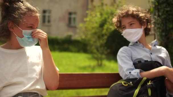 Παιδιά σχολικής ηλικίας με ιατρικές μάσκες κάθονται σε ένα παγκάκι και μετά λένε αντίο, αγγίζοντας ο ένας τον άλλο με τους αγκώνες τους. Ασύρματος αποχαιρετισμός, πίσω στο σχολείο μετά από καραντίνα coronavirus covid-19 — Αρχείο Βίντεο