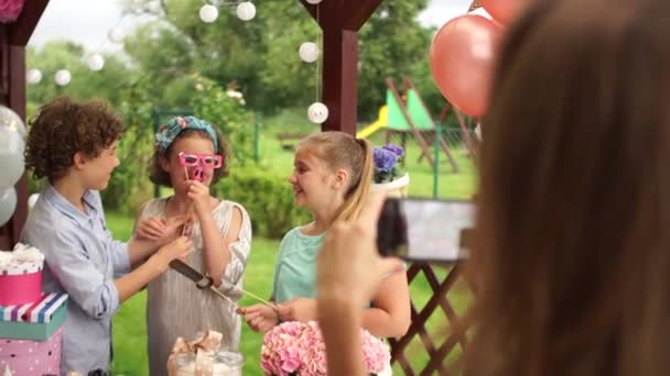 Çocuklar için tatil, doğum günü partisi. Gençler çubuk şekerin yanında fotoğraf çektirip eğleniyorlar. Fotobuthoria bıyığı, şapkası ve büyük dudakları var. — Stok video