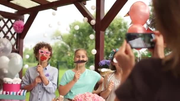 Mama robi zdjęcia na urodzinach dzieci bawiących się rekwizytami fotograficznymi. Photobuthoria na imprezę dla dzieci - papierowe okulary, wąsy i emotikony. Wszystkiego najlepszego z dzieciństwa. — Wideo stockowe