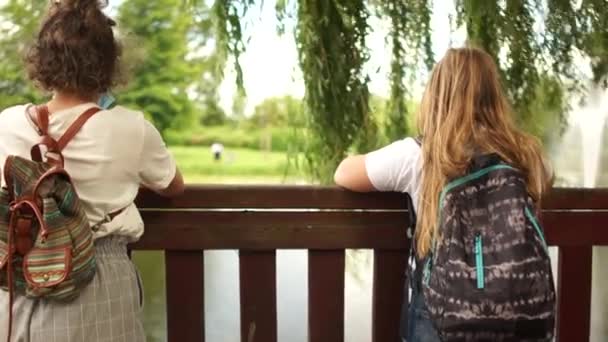 Due studentesse si sono incontrate in un parco vicino al lago. Si mettono le maschere e si salutano senza contatto con il gomito. Studentesse con zaini — Video Stock