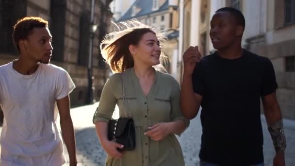 Unga studenter går runt i staden. Två afroamerikanska killar och en vit tjej njuter av en promenad genom staden. Studenter på en promenad, grupp ungdomar mellan raser — Stockvideo