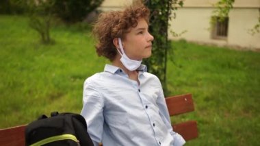 Kıvırcık saçlı okul çocuğu maskesini çıkardı ve rahatça nefes alıyor. Covid-19 Coronavirus salgını sırasında bir çocuk okuldan sonra bankta oturuyor. Karantinadan sonra okula dönüyorum.