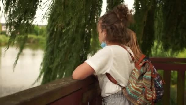 Choolgirl im Park mit ihrem Freund. Die Mädchen haben ihre Masken abgelegt und unterhalten sich, während sie am See stehen. Leben nach der Quarantäne, maskierte Schulkinder. Zurück zur Schule — Stockvideo