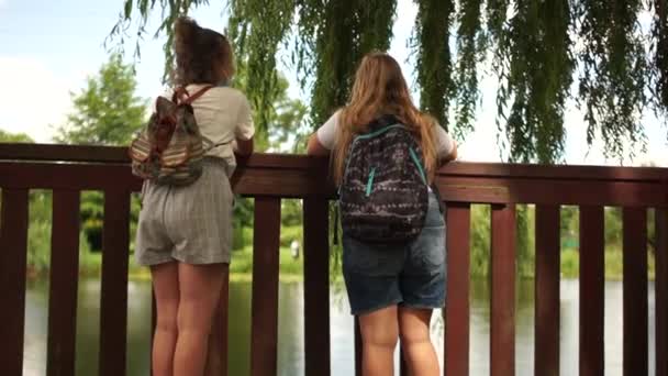 Temassız vedalar, sosyal mesafeler ve okul çocukları. İki liseli kız gölün yakınındaki bir parkta karşılaştı. Maske takıyorlar ve temas etmeden vedalaşıyorlar. Sırt çantalı okul kızları — Stok video