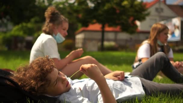 Maszkos iskolás osztálytársak pihennek a füvön. Karantén utáni élet, új normalitás, vissza az iskolába — Stock videók