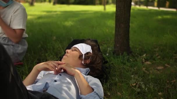 Ein Junge mit Maske ruht nach einem Schultag im Park. Der Schuljunge liegt auf dem Gras und hält einen Grashalm im Mund. Leben nach der Quarantäne. Zurück zur Schule — Stockvideo