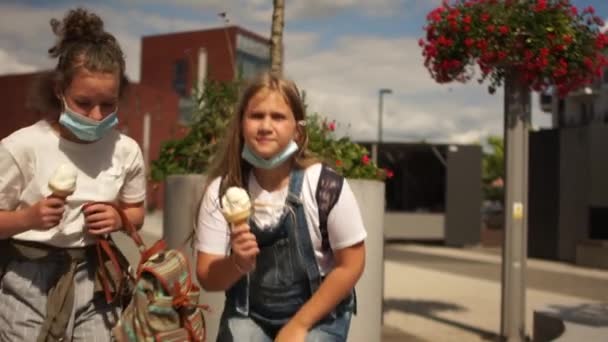 Dos chicas enmascaradas están comiendo helado. Retrato urbano de colegialas, día soleado de verano, coronavirus de cuarentena covid-19 — Vídeos de Stock