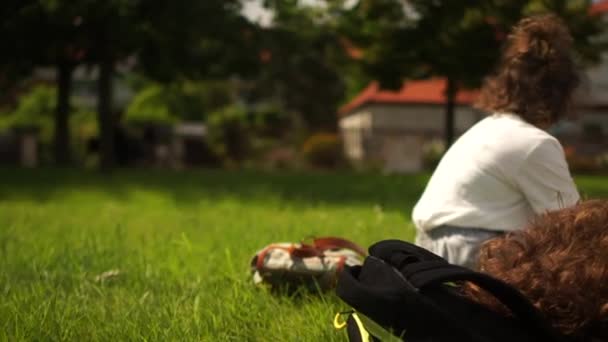 Niños enmascarados con mochilas descansan en el césped del parque. Vida post-cuarentena, nueva normalidad, vuelta a la escuela — Vídeos de Stock