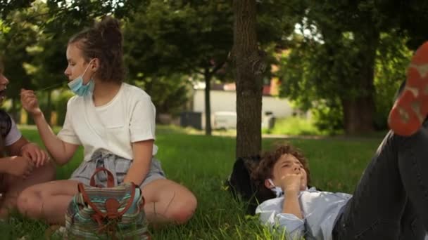 После школьного дня в парке отдыхают дети в масках. Девушка держит травинку во рту. Послекарантинная жизнь. Возвращение в школу — стоковое видео