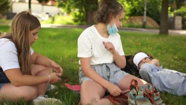 Bezkontaktní pozdravy, nová pravidla. Maskované školáci děti s batohy odpočívají na trávě v parku. Postkaranténní život, nová normalita, návrat do školy — Stock video