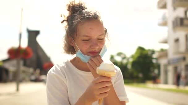 Die schöne, lockige Teenagerin zieht ihre medizinische Schutzmaske ab und isst Eis. Porträt auf einer Straße in der Stadt. Sommer während der COVID-19-Coronavirus-Pandemie — Stockvideo