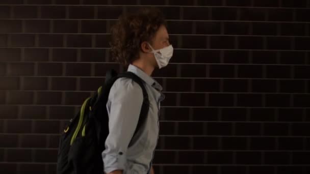 Um estudante de cabelos encaracolados com uma máscara protectora médica vai para a escola. Um menino de camisa azul carrega uma mochila nas costas, uma criança caminha ao longo de uma passagem subterrânea. Voltar para a escola covid — Vídeo de Stock