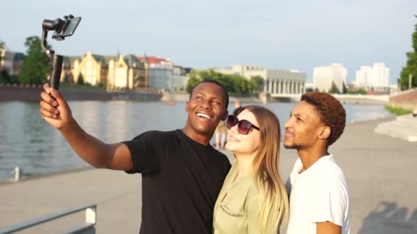 Tre unge studerende tager selfies ved hjælp af en smartphone og selfie stick. To sorte mænd og hvid kvinde har det sjovt på byens gader, rejser koncept – Stock-video