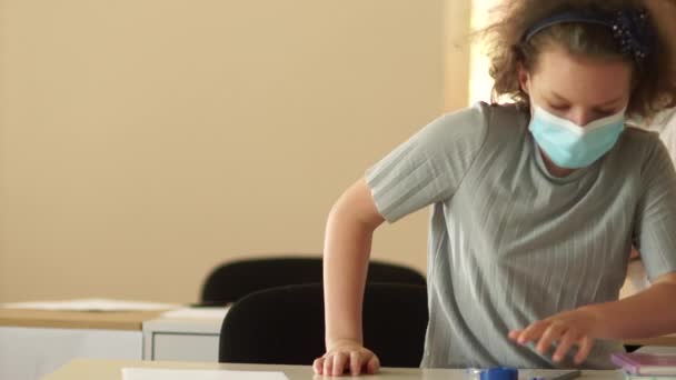 De volta à escola depois da quarentena do coronavírus covid-19. Reabrir escolas. Uma menina com uma máscara protetora sentou-se em sua mesa, colocou seus óculos e abriu um caderno. Observe cuidadosamente — Vídeo de Stock