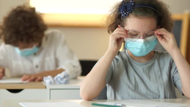 Curly schoolmeisje draagt een bril. Kinderen gezichtsmasker. Terug naar school covid. Kinderen met beschermende medische maskers zitten aan hun bureau op school — Stockvideo