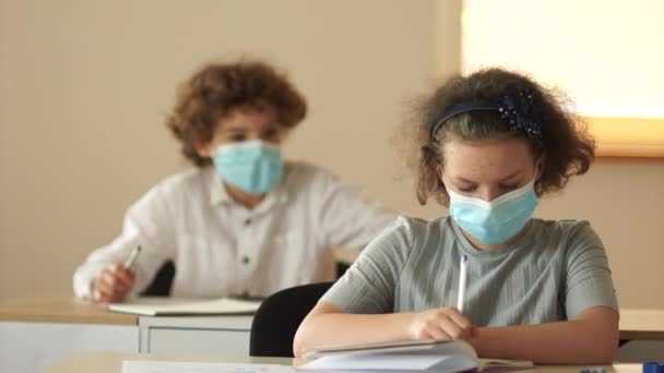Compañeros de clase con máscaras médicas protectoras, un niño y una niña están sentados en sus escritorios en la escuela. El chico le pide a la chica un papel. Regreso a la escuela — Vídeo de stock