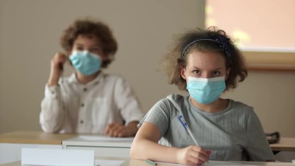 Çocuklar maske takıyor. Okuluna geri dön. Koruyucu tıp maskeli çocuklar okulda masalarında otururlar. Kıvırcık kız liseli — Stok video