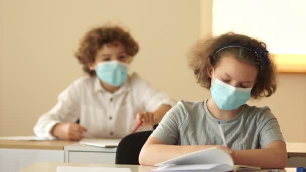 Zurück zur Schule. Mitschüler in medizinischen Schutzmasken, ein Junge und ein Mädchen sitzen in der Schule am Schreibtisch — Stockvideo