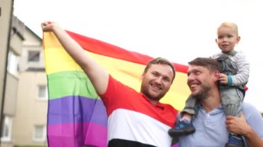 Küçük çocuklu homoseksüel çift LGBT bayrağı taşıyor. Gökkuşağı bayrağı, açık hava portresi, iki adam ve bir çocuk, evlatlık çocuklar, evlat edinme prosedürü