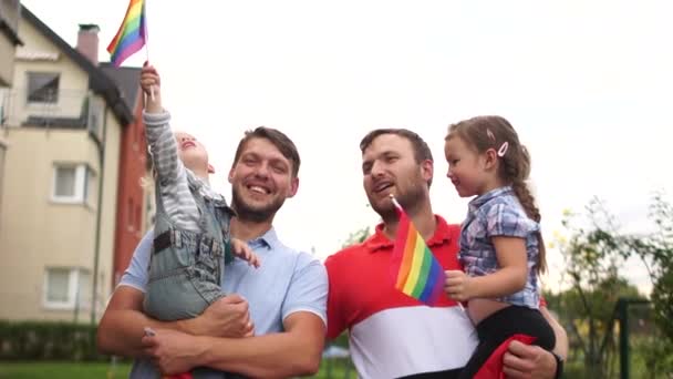 Adoptované děti. Gay pár. Dva muži s dítětem se políbí. Homosexuální rodina se dvěma dětmi držícími vlajky LGBT na pozadí domu a oblohy — Stock video