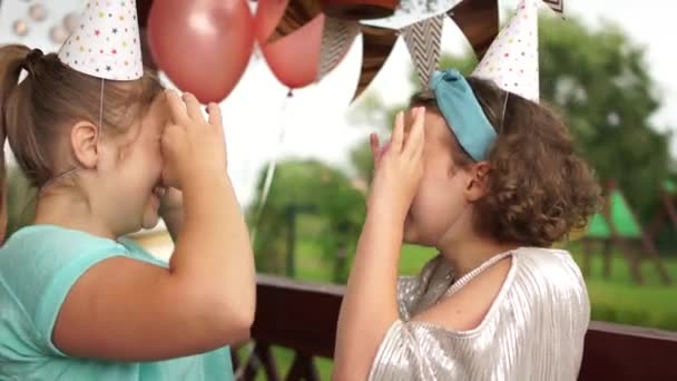 Szalone wesołe dziewczyny w urodzinowych kapeluszach uśmiechnięte, bawiące się i patrzące przez dwa różowe ciasteczka na ich oczach. Słodycze. Portret na świeżym powietrzu, impreza dla dzieci, wszystkiego najlepszego — Wideo stockowe