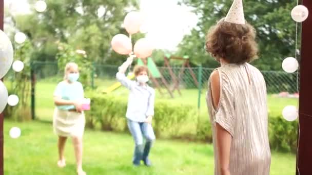 Fête d'anniversaire pendant la quarantaine de covide-19. Une fille d'anniversaire portant un masque accueille les invités à sa fête en plein air. Des enfants masqués lui donnent des cadeaux et des ballons. Distance sociale — Video