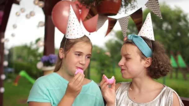 Δύο έφηβες με χάρτινα καπέλα τρώνε ροζ μπισκότα. Παιδικό πάρτι γενεθλίων, γλυκό τραπέζι, χαρούμενα παιδιά — Αρχείο Βίντεο