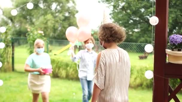 Une fille d'anniversaire portant un masque accueille les invités à sa fête en plein air. Des enfants masqués lui donnent des cadeaux et des ballons. Distance sociale. Fête d'anniversaire pendant la quarantaine de covid-19 — Video