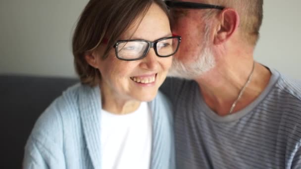 Feliz pareja de ancianos enamorados. Viejo susurrando secreto a su esposa. Hombres y mujeres de pelo gris están sentados en el sofá. Mujer lleva gafas y ríe alegremente — Vídeos de Stock